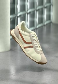 Baskets crème et marron clair à lacets avec semelle rainurée et marquage "BOSS" sur le côté, présentées en suspension au-dessus d'un sol gris.