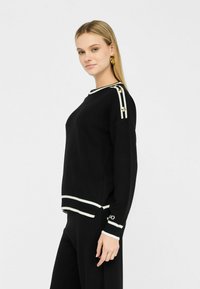 Zwarte pullover sweater met witte gestreepte accenten op de halslijn en de zoom. Voorzien van gouden hardware knopen op de schouders en een ribbelstructuur.