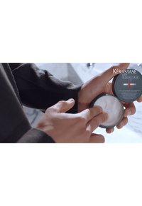 Crema para el cabello de Kérastase en un envase circular oscuro. Se está aplicando una textura de crema blanca con los dedos. El envase presenta etiquetado en gris y rojo.