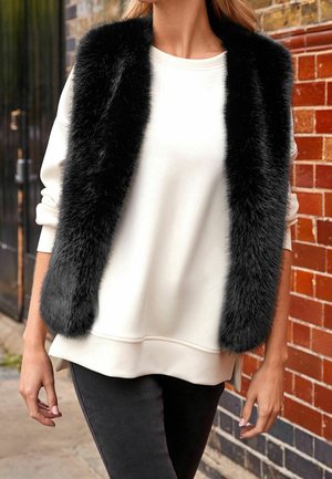 Femme portant une veste en fausse fourrure noire sur un pull blanc à manches longues, debout contre un mur en briques à l'extérieur.