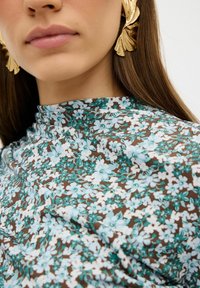 Blumenmusterstoff in Hellblau und Weiß auf einem braunen Hintergrund. Besitzt einen hohen Ausschnitt und eine glatte Textur, kombiniert mit dekorativen goldenen Ohrringen.