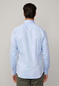 Camicia a maniche lunghe blu chiaro con collo classico, cucitura posteriore dritta e polsini con bottoni. Il materiale appare liscio con una vestibilità su misura.