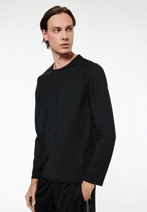 Młody mężczyzna o średniej długości włosach, ubrany w czarną koszulę z długim rękawem i czarne spodnie, patrzący w lewo z jedną ręką w kieszeni.