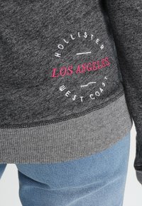 Grå sweatshirt med en texturerad yta, med en rosa och vit logotyptext: "HOLLISTER LOS ANGELES WEST COAST," och en randig nederkant.