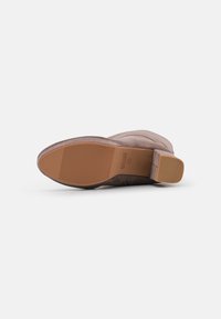 Botín de ante marrón claro con una superficie texturizada, punta redondeada y un tacón cuadrado de altura media. Suela de goma con estrías verticales.