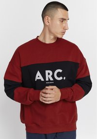 Rød og svart hettegenser med lange ermer, med et iøynefallende hvitt grafisk trykk "ARC." over brystet. Myk tekstur og avslappet passform.