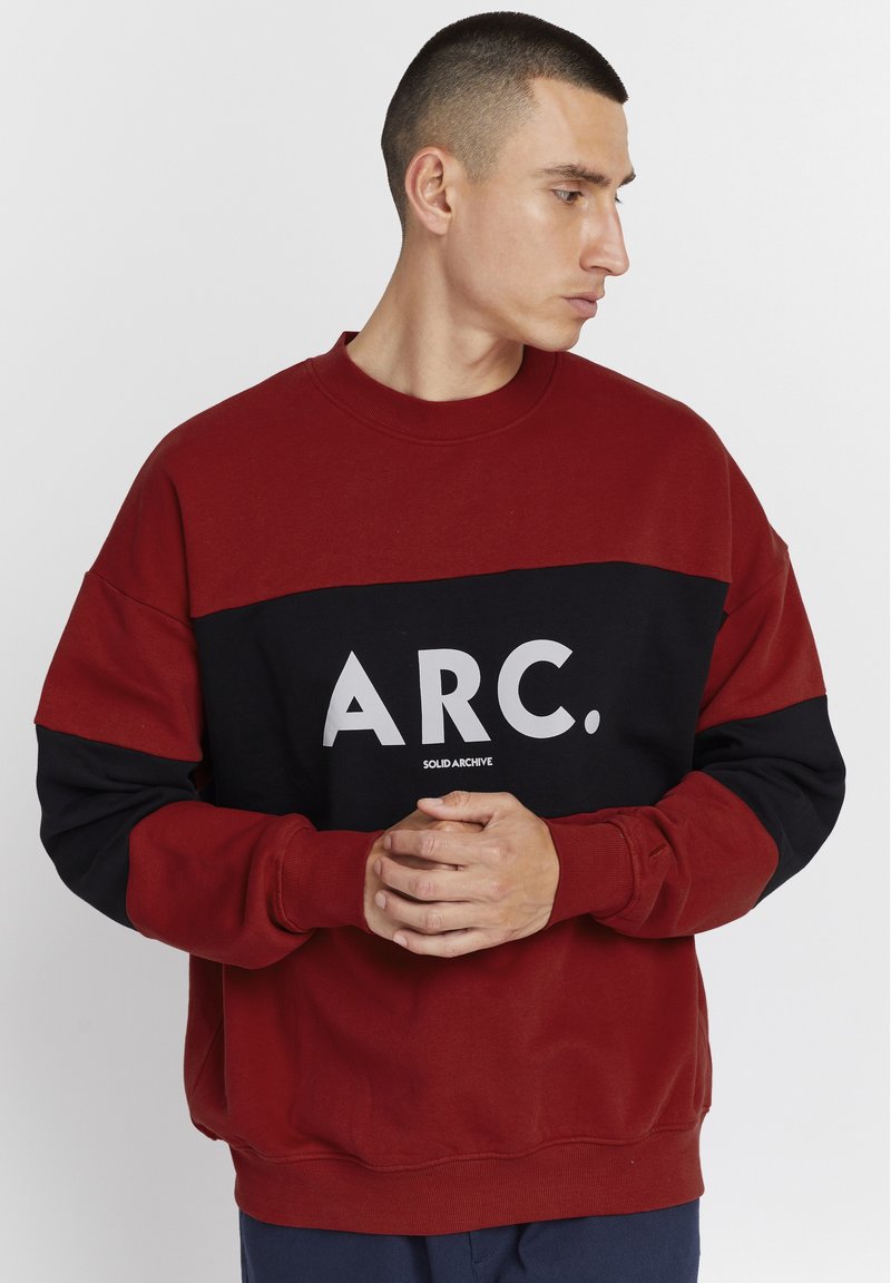 Rød og svart hettegenser med lange ermer, med et iøynefallende hvitt grafisk trykk "ARC." over brystet. Myk tekstur og avslappet passform.