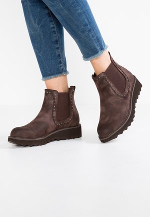 Bottines en daim marron avec bordure cloutée et semelles épaisses nervurées portées avec un jean bleu à ourlet effiloché sur fond blanc uni.