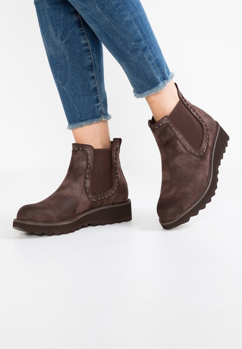 Bottines en daim marron avec bordure cloutée et semelles épaisses nervurées portées avec un jean bleu à ourlet effiloché sur fond blanc uni.