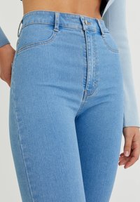 Korkeavyötäröiset siniset farkut, joissa on sileä pinta, varustettuna napilla ja vetoketjulla, sekä standardin viiden taskun muotoilu.