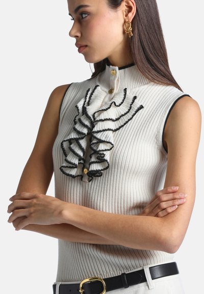 Femme aux longs cheveux foncés portant un haut blanc côtelé sans manches avec des volants bordés de noir et des boutons dorés, ceinture noire et boucles d'oreilles dorées, bras croisés.