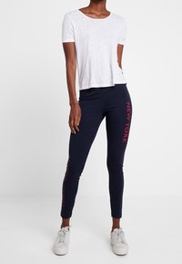 T-shirt blanc à manches courtes associé à un legging marine avec du texte rouge "NEW YORK" sur le côté. Les chaussures sont des baskets blanches.