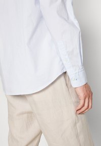 Hellblaues Hemd mit entspannter Passform, aus glattem Baumwollmaterial und abgerundetem Saum, kombiniert mit beigen Leinenhosen.
