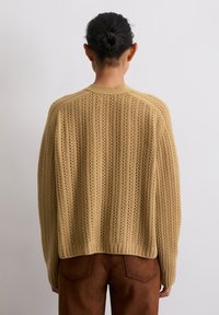 Beiger Strickpulli mit vertikalem Rippmuster, Durchbrüchen, Rundhalsausschnitt und tief sitzenden Schultern, kombiniert mit braunen Wildlederhosen.
