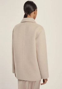 Cappotto di lana beige con colletto a revers, maniche lunghe e vestibilità rilassata. Presenta una costruzione semplice e una finitura testurizzata, visto da dietro.