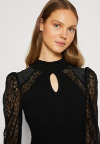 Zwarte gebreide top met lange kant mouwen, met een keyhole halslijn. Het kant heeft bloemmotieven en doorschijnende accenten op de schouders.