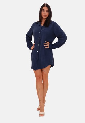 Femme aux cheveux foncés portant une robe chemise bleu marine à manches longues boutonnée et des talons ouverts beige, debout les mains sur les hanches.