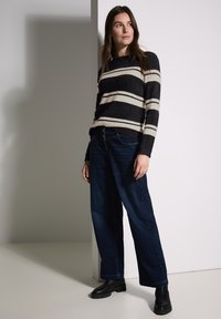 Pull rayé en noir et crème, tissu texturé, associé à un jean large bleu foncé. Porté avec des bottines noires.