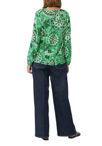 Grüne gemusterte Bluse mit langen Ärmeln, kombiniert mit dunkelblauen Hosen mit weitem Bein. Die Bluse zeichnet sich durch aufwendige Blumenmuster und einen leichten Glanz aus.