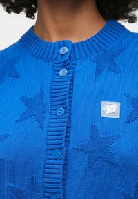 Plavi pleteni kardigan s rebrastim ovratnikom i pet usklađenih dugmadi. Teksture zvjezdastih uzoraka i kvadratna oznaka na prsima.