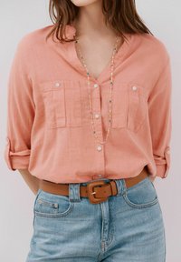 Camicia button-up rosa floreale con due tasche sul petto, maniche arrotolate, abbinata a jeans azzurri chiari e una cintura color crema, ornata con una collana di perline colorate.