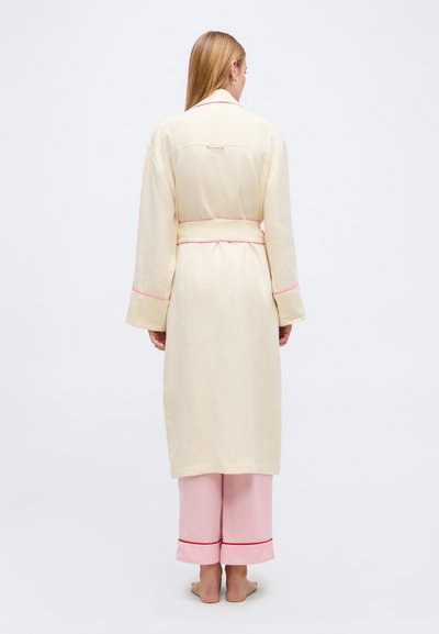 Γυναίκα που στέκεται ξυπόλητη με την πλάτη, φοράει μια κρεμ μακριά ρόμπα με ροζ επένδυση και αντίστοιχο ροζ παντελόνι πιτζάμας με κόκκινο περίγραμμα.