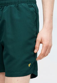 Mehe käsi puhkab tumeroheliste lühikeste pükste tasku lähedal, millel on elastne vöökoht ja vasakul jalal väike kollane kiililinnu logo.