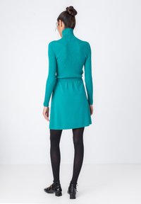 Turkizna rebrasta turtleneck pulover, združen s turkizno razširjeno krilo, nosi se z črnimi najlonkami in gležnjarji z zlatimi detajli.