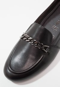 Mocassin en cuir noir avec un détail de chaîne argentée sur le dessus, présentant une texture lisse et un design à bout arrondi.