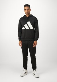 Moletom preto com logótipo triangular branco, combinado com calças de moletão pretas. O modelo usa sapatilhas brancas com riscas pretas. Tecido suave, design casual.