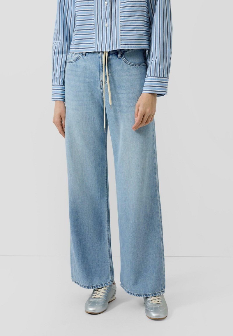 Osoba ubrana w jasnoniebieskie szerokie jeansy z wiązaniem w talii, niebieską koszulę w paski oraz srebrne trampki na jednolitym szarym tle.