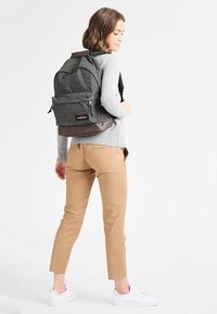 Mochila de tela gris con una superficie texturizada, fondo marrón en contraste, dos bolsillos frontales y correas ajustables, llevada con un atuendo casual.