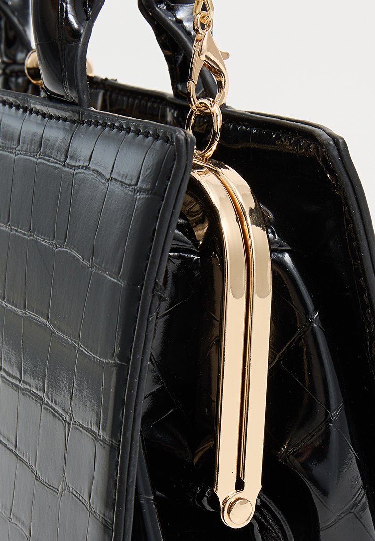 Sac à main en cuir texturé noir avec fermoir en métal doré et petite poignée accrochée sur un fond uni.