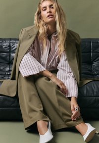 Olīvu zaļš palielināts bleizeris virs rozā un baltām svītrām krekliem, apvienots ar platām olīvu krāsas biksēm un baltām slēgtajām kurpēm ar papēžiem.