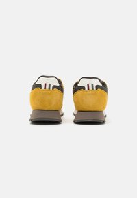 Pantofi sport cu un spate din suede galben, parte superioară din material textil maro, detaliu alb pe călcâi cu dungi roșii și albastre, și talpă externă din cauciuc gri.