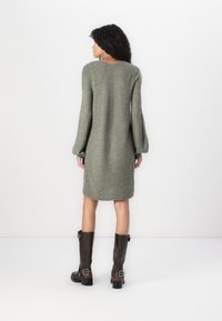 Robe en tricot verte avec de longues manches et un col rond. Présente une coupe décontractée, un ourlet côtelé, et est associée à des bottes marron montantes.