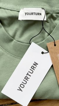 Světlounce zelené bavlněné tričko s kulatým výstřihem a viditelným švem. Připevněné štítky zahrnují bílou nálepku s nápisem "YOURTURN" a hnědý štítek.