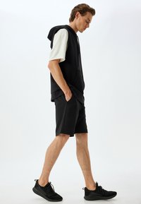 Sudadera sin mangas negra sobre una camiseta clara, combinada con pantalones cortos negros y zapatillas negras. Diseño simple con bolsillos.