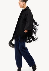 Femme marchant dans une veste noire à franges, un jean large foncé, des bottines noires, de grandes boucles d'oreilles en argent, portant un sac à main noir clouté.