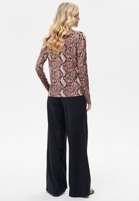 Top met lange mouwen met een roze en bruin slangenprintpatroon, gecombineerd met zwarte broek met brede pijpen. Model getoond van achteren.