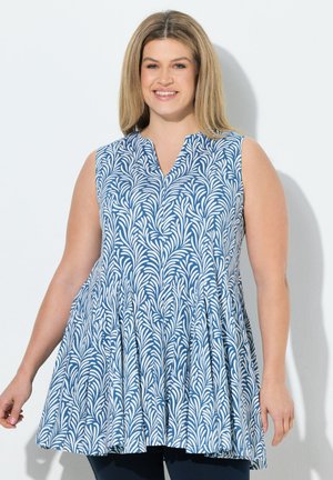 Smilende kvinde iført ærmeløs tunika i blå og hvid med bladlignende mønster, stående foran en ensartet lys baggrund.