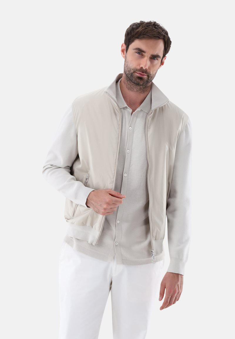 Hellbeige gepolsterte Weste mit hohem Kragen über einem hellgrauen Poloshirt. Sie verfügt über seitliche Reißverschlusstaschen sowie ein geripptes Saum- und Manschettendesign.