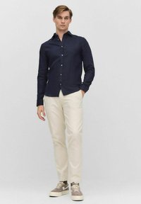 Marineblauw overhemd met lange mouwen, gecombineerd met crèmekleurige broeken. Model draagt grijze sneakers met witte zolen en een casual houding.