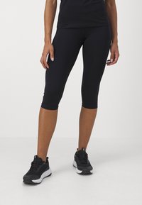 Fekete capri leggings, szabott dizájnnal, rugalmas anyagból, fekete sportcipőkkel kombinálva, amelyek texturált felsőrésszel és fehér talppal rendelkeznek.