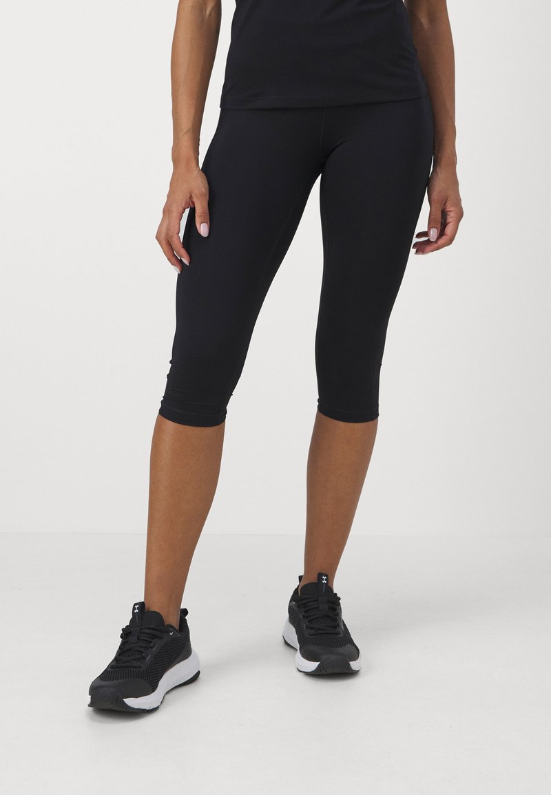 Fekete capri leggings, szabott dizájnnal, rugalmas anyagból, fekete sportcipőkkel kombinálva, amelyek texturált felsőrésszel és fehér talppal rendelkeznek.