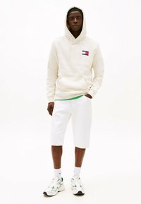 Felpa crema con tasca frontale e toppa con logo, abbinata a pantaloni corti bianchi e scarpe da ginnastica con dettagli verdi. Sfondo bianco.