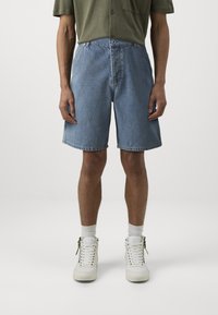 Shorts en jean bleu clair, longueur genou avec une coupe décontractée, dotés d'une fermeture à bouton à l'avant et de poches classiques. Portés avec des baskets blanches.
