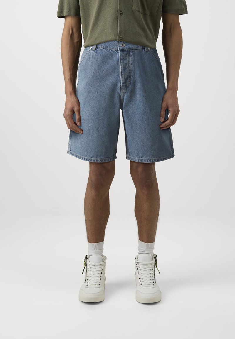 Denimshorts i lys blå, kne-lange med en avslappet passform, med knappelukking foran og standard lommer. Brukt med hvite joggesko.