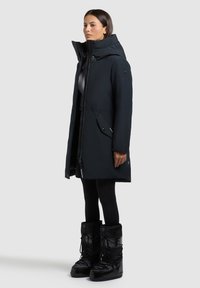 Sort isoleret parka med høj krave, skjult lynlås, sidelommer og mat finish, båret over sorte leggings og blanke sorte støvler.