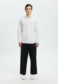 Bílá košile s button-up límcem a jemným pruhováním, v kombinaci s volnými černými kalhotami a bílými teniskami, stojící na světle šedém pozadí.